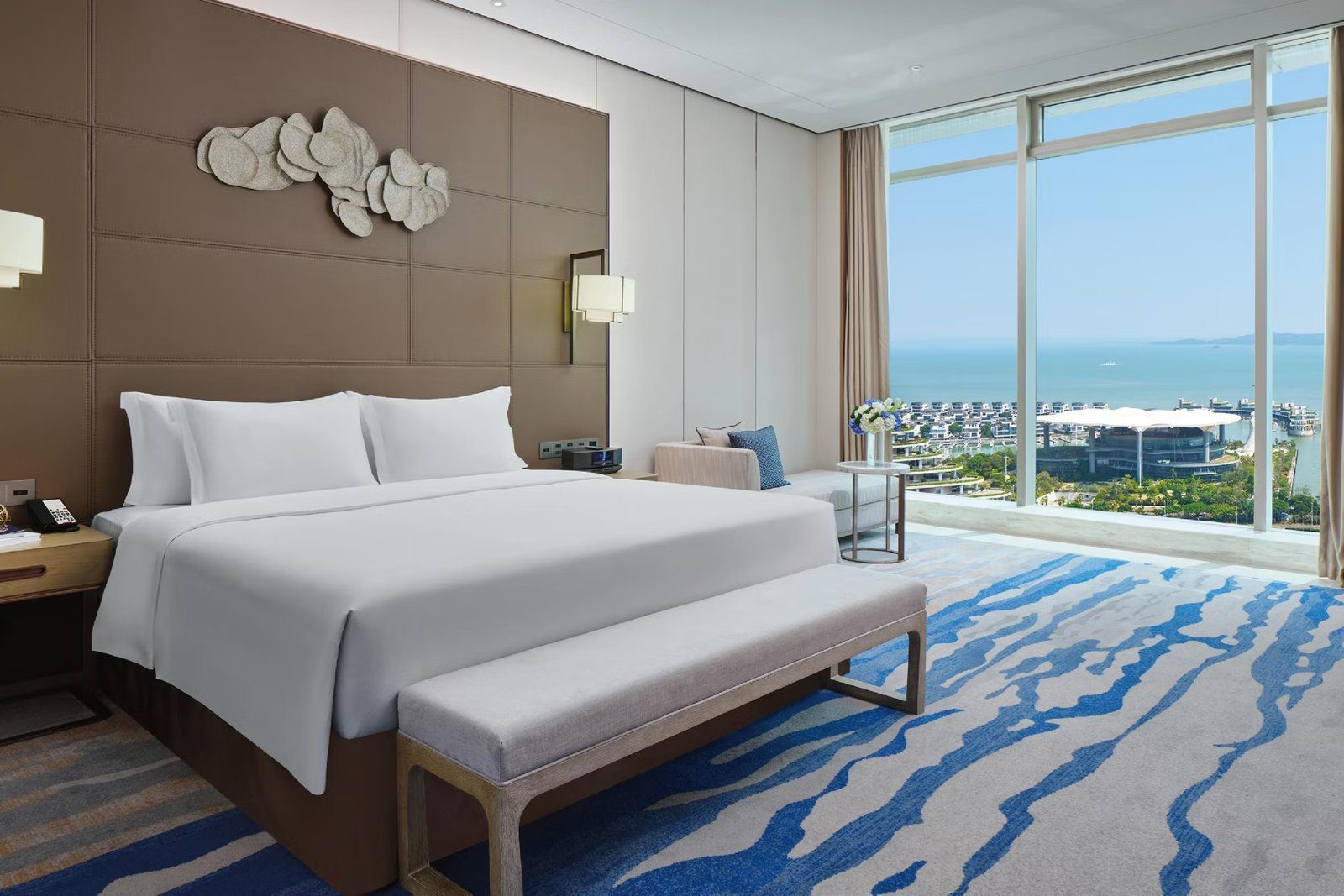 廈門海景洲際酒店的海景客房，白色寢具的大床靠著皮革格紋床頭牆，床尾擺淺灰色長凳，地毯是藍白水波紋，右側整面落地窗外是海面、海岸綠地與白色棚頂建築
