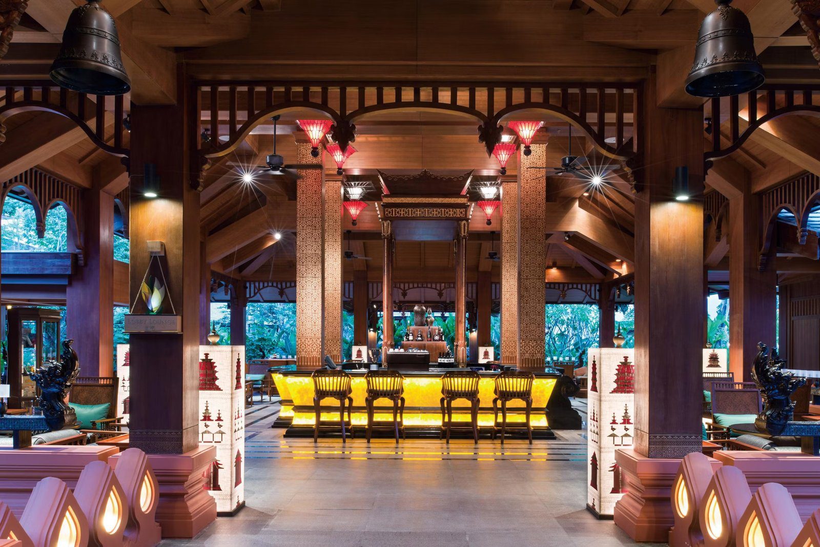 傣式木構大廳裡的吧台，吧台面透出黃色燈光，前方立柱掛著 LOBBY LOUNGE 牌子，上方垂著紅色燈罩與銅鐘