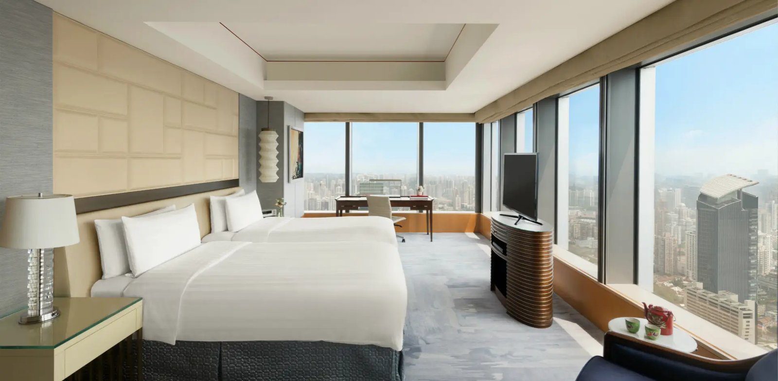 上海靜安香格里拉雙床客房，兩張單人床靠著米色格紋床頭牆，轉角落地窗前是書桌與電視櫃，窗外是市區高樓