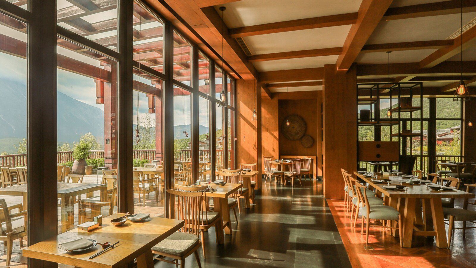 麗江金茂璞修雪山酒店 The Lodge 餐廳的用餐區，木樑下排著原木桌椅，左側整面落地窗外是戶外露台座位與山景