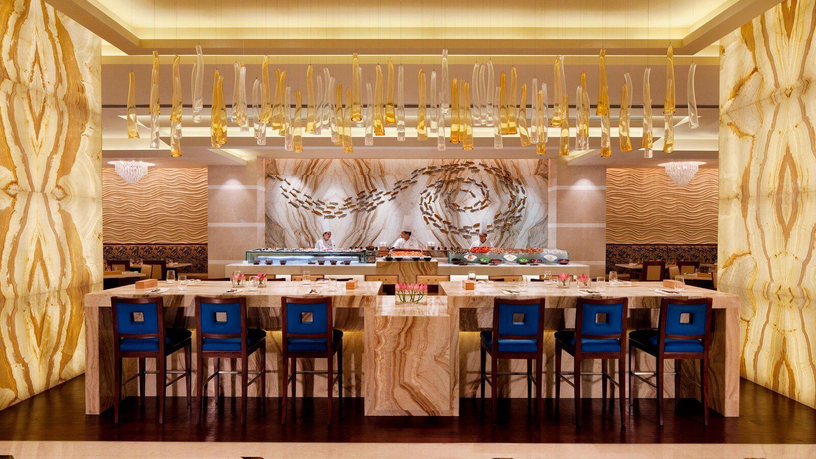 杭州 JW 萬豪酒店 Asia Bistro 的大理石吧檯與海鮮壽司烹調台，後方三名廚師在崗，天花懸著琥珀色玻璃裝置