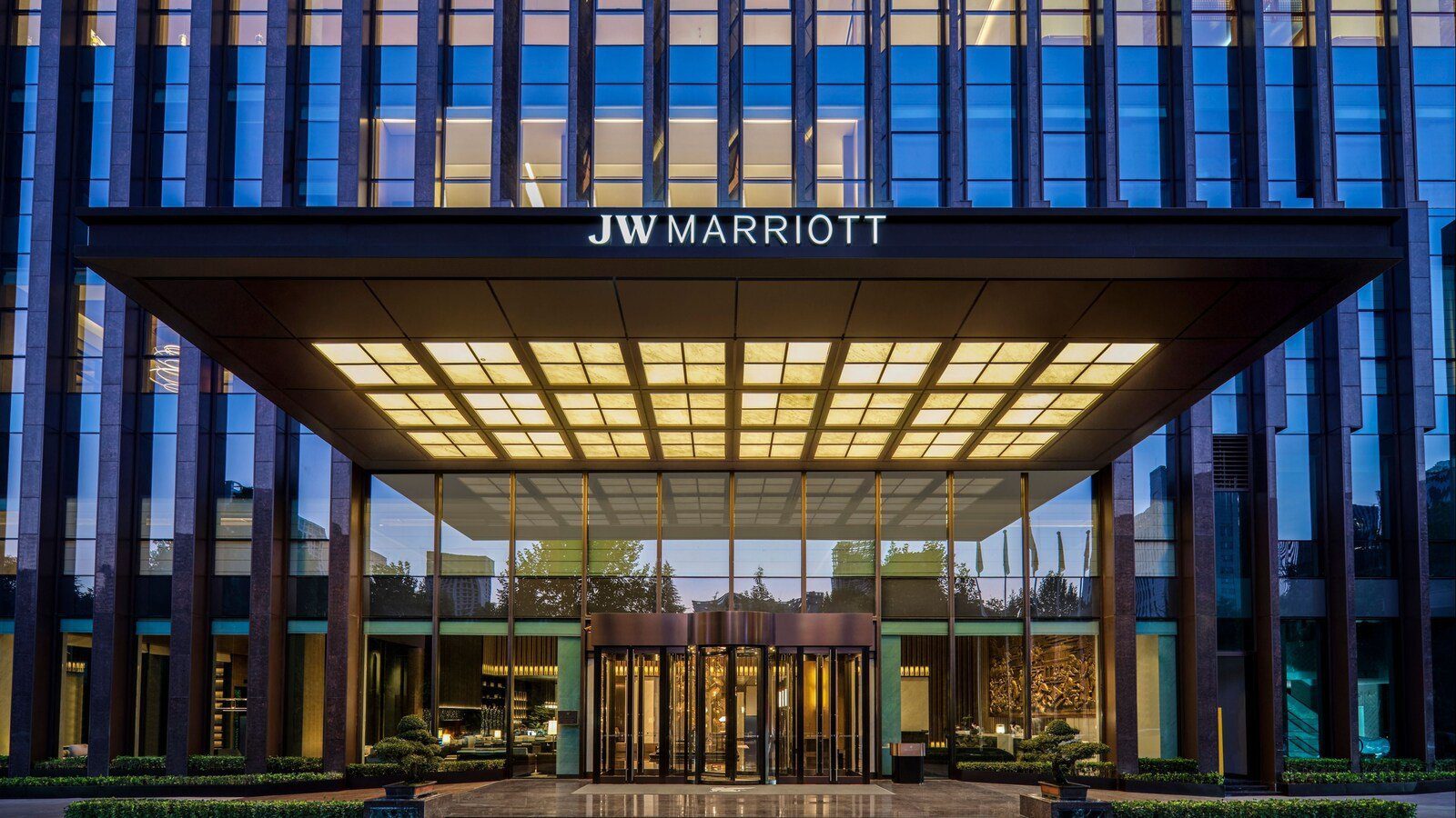 西安高新 JW 萬豪酒店入口：傍晚時分，深色直條立面下方伸出大型雨遮，雨遮上有 JW MARRIOTT 字樣，下方亮著方格燈光，中央是旋轉門，兩側擺著盆景