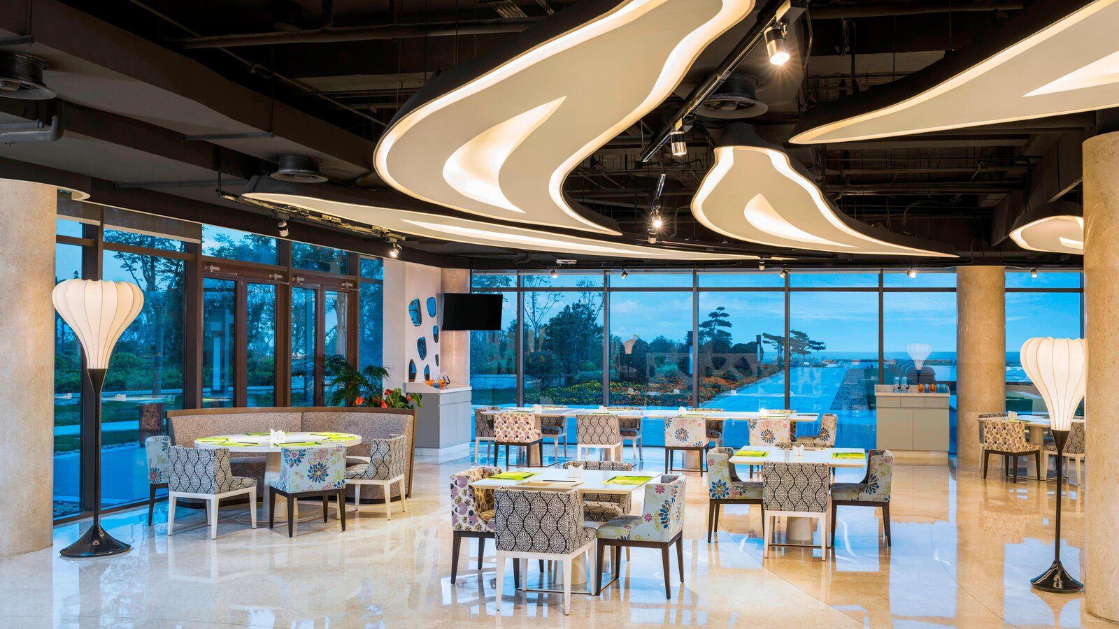 青島西海岸隆和艾美度假酒店新食譜全日餐廳，波浪形燈飾下擺著花紋餐椅，落地窗外是庭園與海