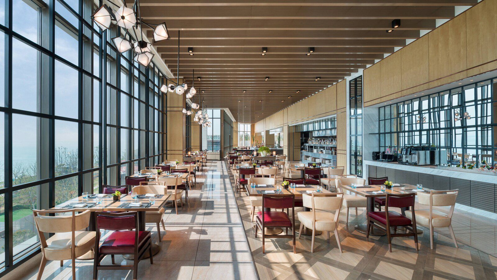 蘇州蘇州灣艾美酒店全日餐廳的長廊式用餐區：整面格狀落地窗採光、木肋條天花與幾何吊燈，酒紅與米色餐椅配木桌，右側是長條備餐吧檯