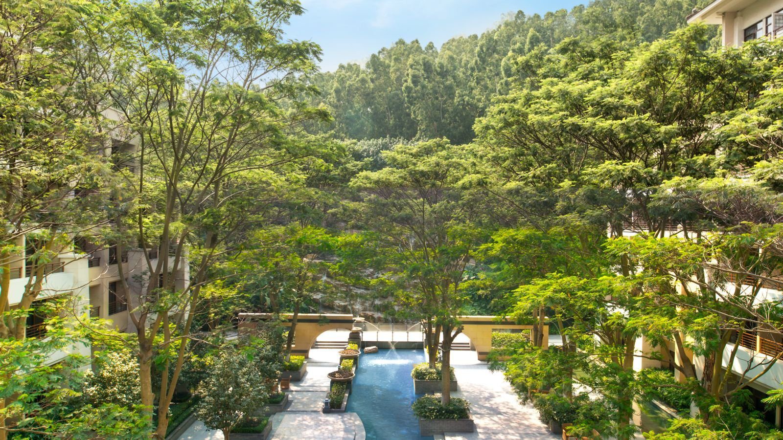 廈門艾美酒店的中庭花園：高大的綠樹圍著一道藍色水景與步道，兩側是客房樓的陽台，遠處是山坡林木與拱門造型的矮牆