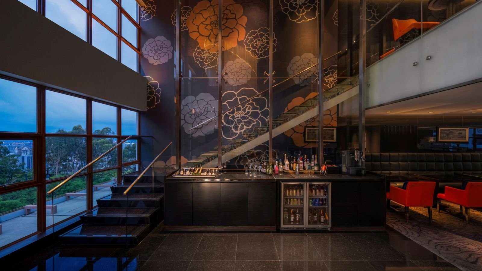 廈門艾美酒店行政酒廊：挑高空間裡的深色吧檯與酒櫃，後方是花卉圖案牆面與玻璃樓梯，左側大片木框窗外是山林與傍晚的城市，右側有紅色扶手椅