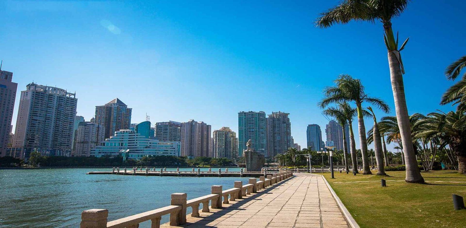 晴天的湖畔步道，石欄杆沿著湖岸延伸，右側是草坪與一排高大的棕櫚樹，湖中有一座石雕像，對岸是成排的住宅與辦公高樓