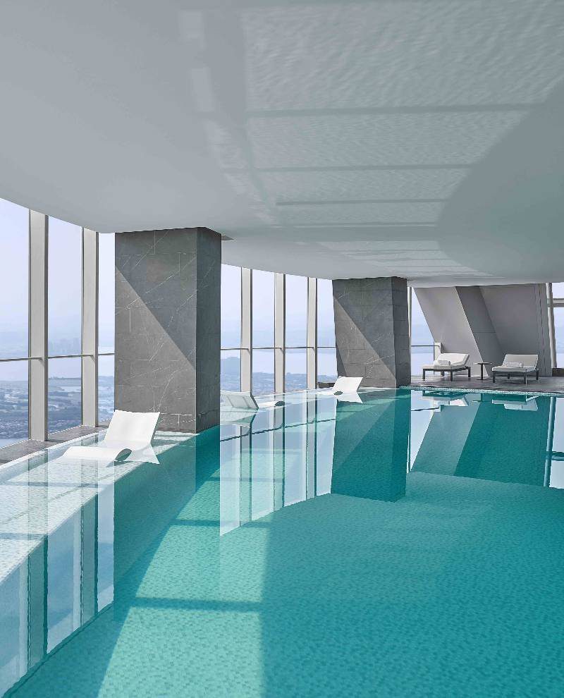 蘇州尼依格羅酒店 Sky Pool 室內泳池，青綠色池水映著灰色石材方柱，一側落地玻璃帷幕向外傾斜、可看城市與湖面，池中設白色躺椅