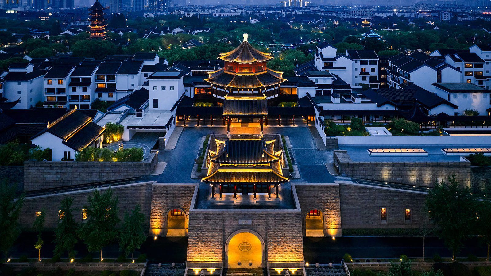 夜間空拍盤門風景區（酒店周邊景點）的城樓與磚砌城牆，燈光打亮重簷屋頂，左後方遠處可見一座亮燈的古塔與蘇州古城民居群