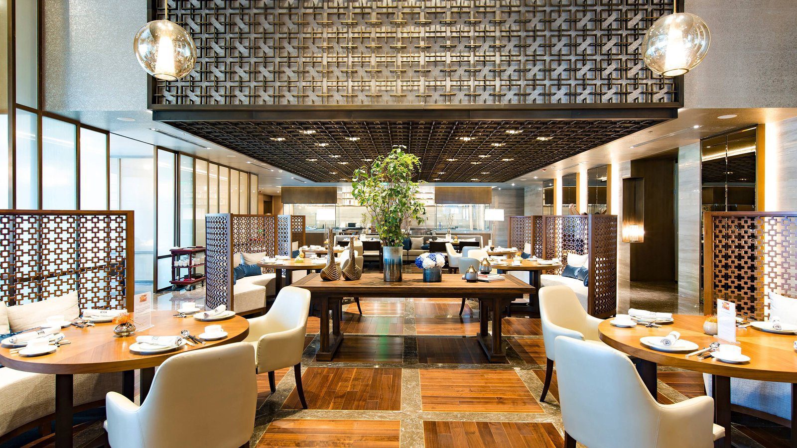 北京富力萬麗酒店雅苑中餐廳大廳，金屬格柵天花與木格屏風隔出座位區，圓桌搭配白色扶手椅，中央長桌擺著綠植