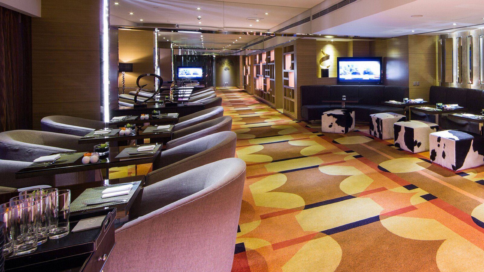 成都首座萬麗酒店 Club 酒廊：左側為成排灰色圓弧扶手椅與深色方桌，右側是皮革長沙發與牛毛紋方凳，地面鋪橘黃色幾何圖樣地毯