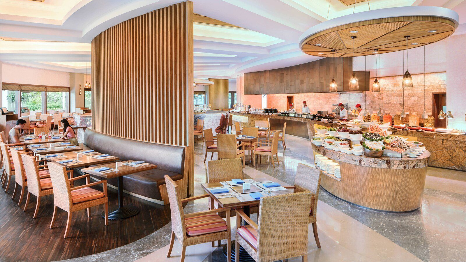 萬豪咖啡廳的用餐區，藤編椅與木桌排開，右側圓弧餐檯擺滿冷盤與海鮮，後方廚師站在開放式廚房前