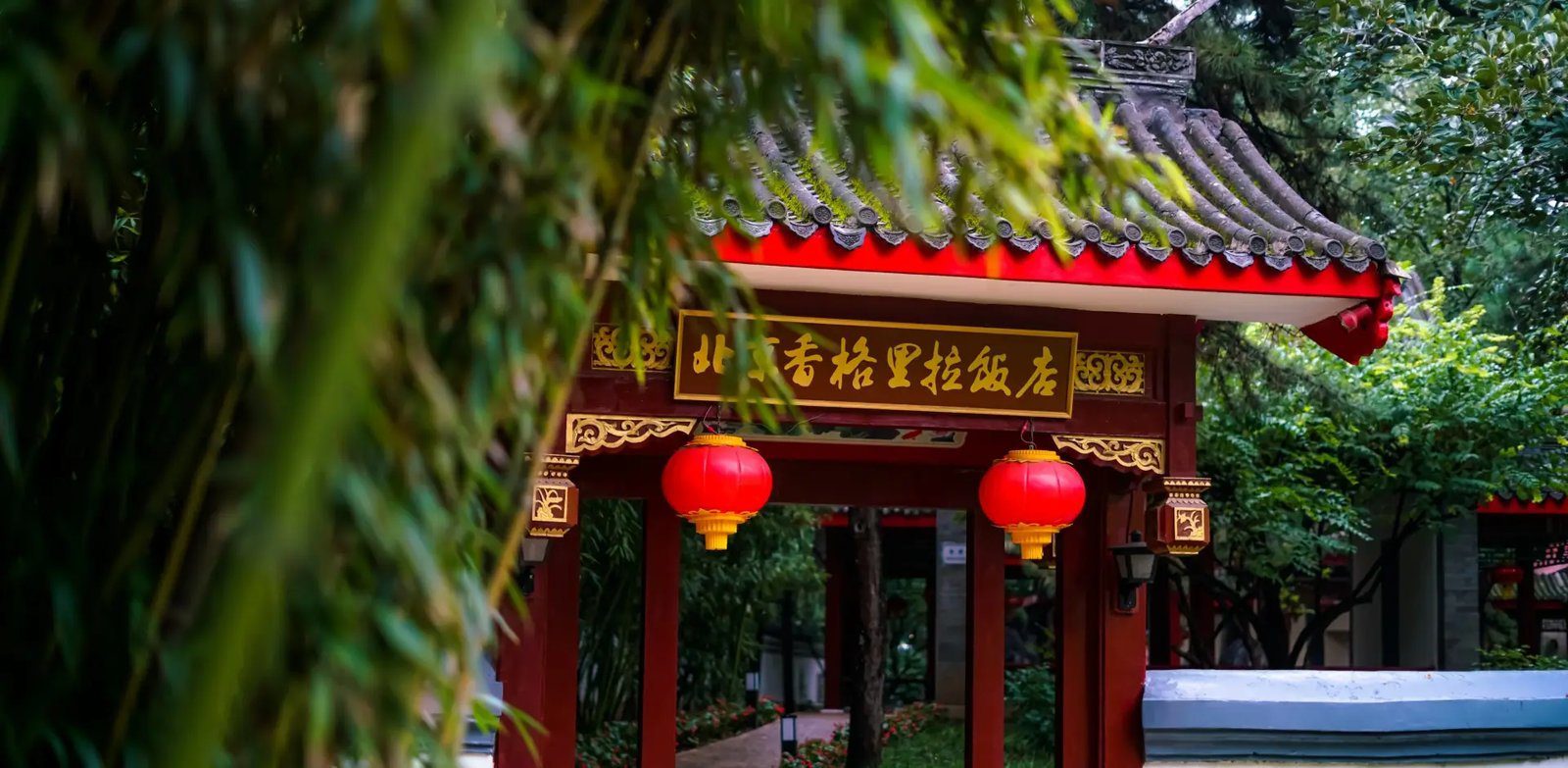 北京香格里拉飯店花園裡的紅柱牌樓，匾額寫著北京香格里拉飯店，簷下掛著兩盞紅燈籠，前景是竹葉