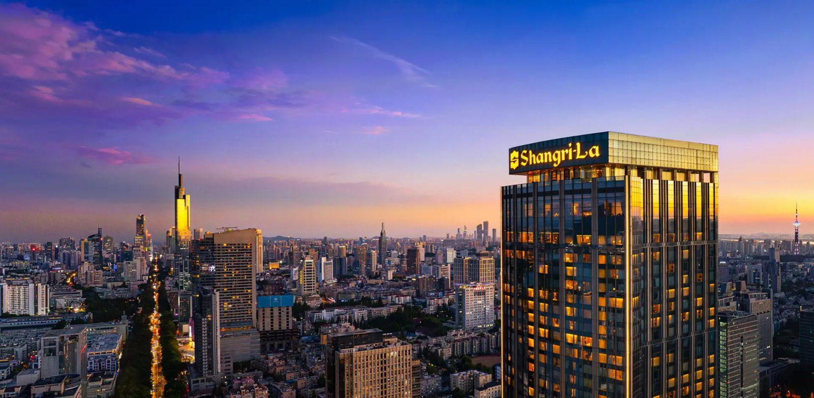 南京香格里拉大酒店黃昏外觀，塔樓頂端亮著 Shangri-La 招牌，左側是亮燈的市區街道與紫峰大廈
