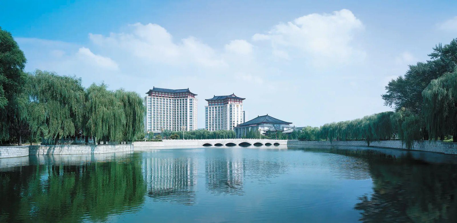 曲阜香格里拉大酒店遠景，兩棟飛簷塔樓立在柳樹夾岸的河面後方，前方有一座多孔石橋