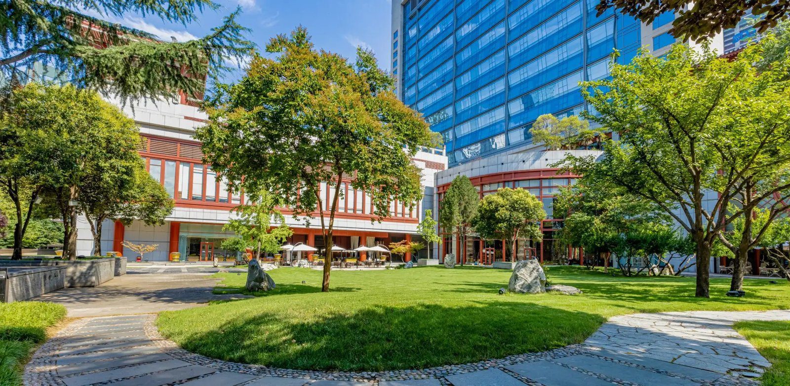 晴天的西安香格里拉花園，弧形石板步道圍著大片草坪與景觀石，四周是綠樹，後方是紅色窗框的裙樓、白色陽傘下的戶外座位與藍色玻璃塔樓