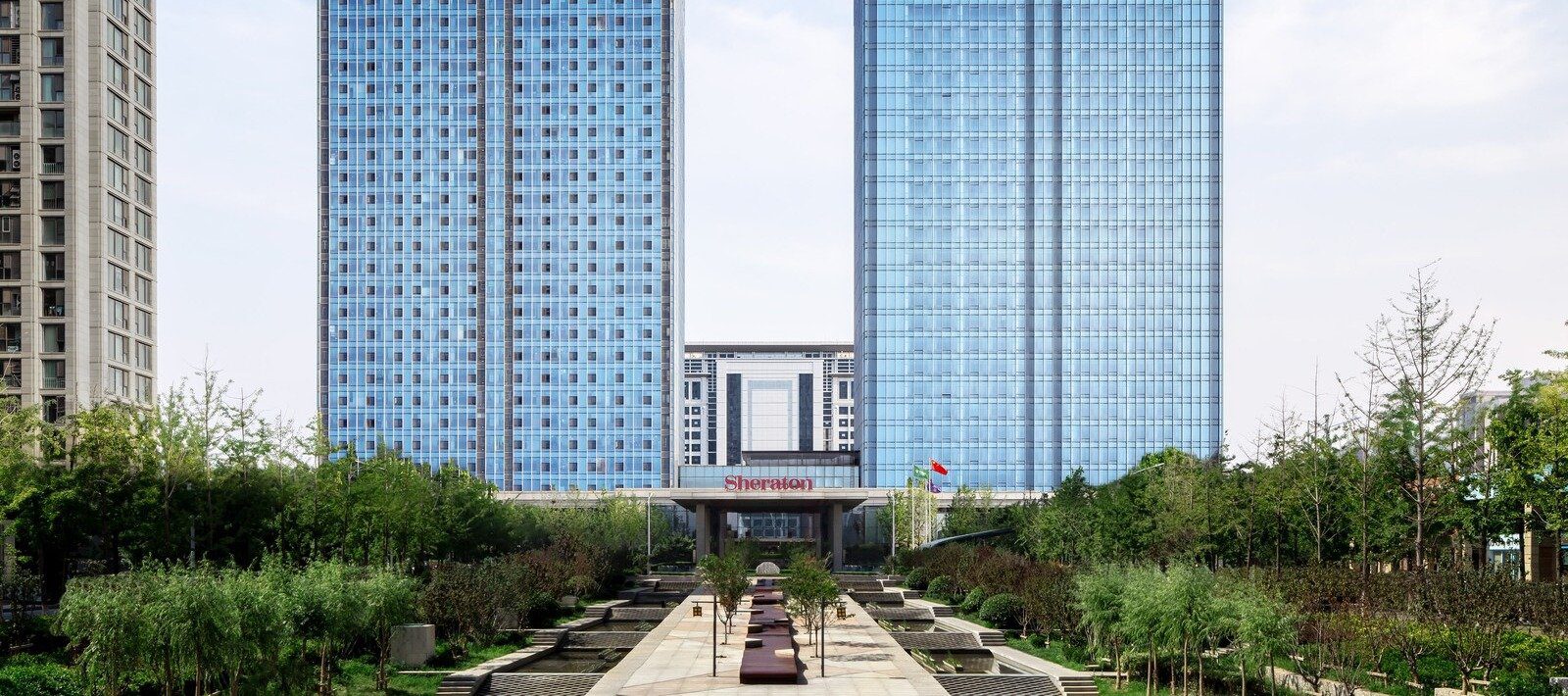 青島膠州綠城喜來登酒店白天外觀，玻璃高樓前是 Sheraton 招牌的入口，前方是種滿樹的水景步道