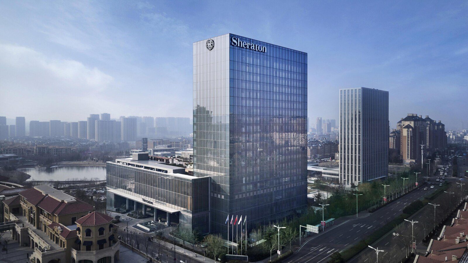 青島李滄綠城喜來登酒店白天外觀，玻璃帷幕塔樓頂端是 Sheraton 招牌，左側是低矮的裙樓與湖面