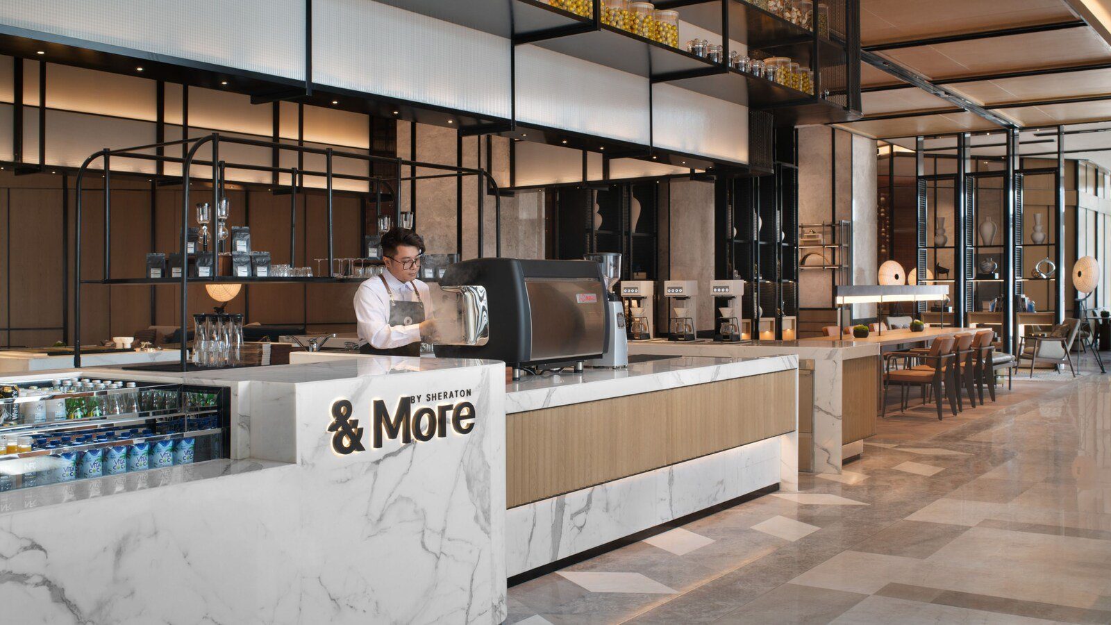 大堂的 &More by Sheraton 咖啡吧,白色大理石吧檯後方有咖啡師操作咖啡機,右側是長桌座位區
