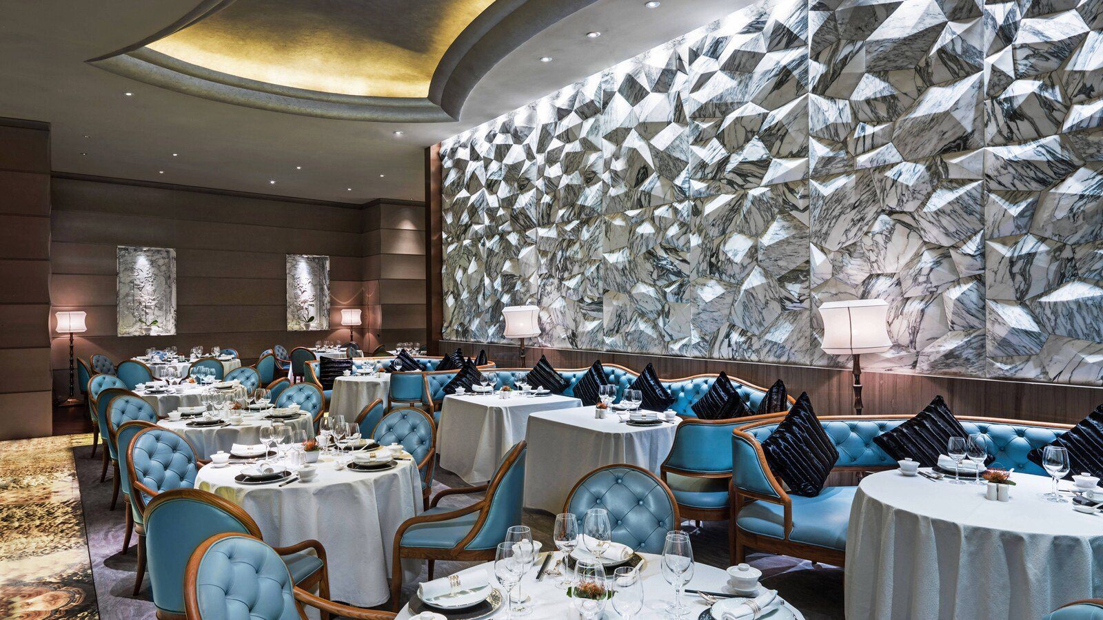 成都瑞吉酒店宴庭中餐廳的主餐區：鋪白桌布的圓桌配水藍色絨布扶手椅與卡座，整面牆為立體幾何浮雕大理石