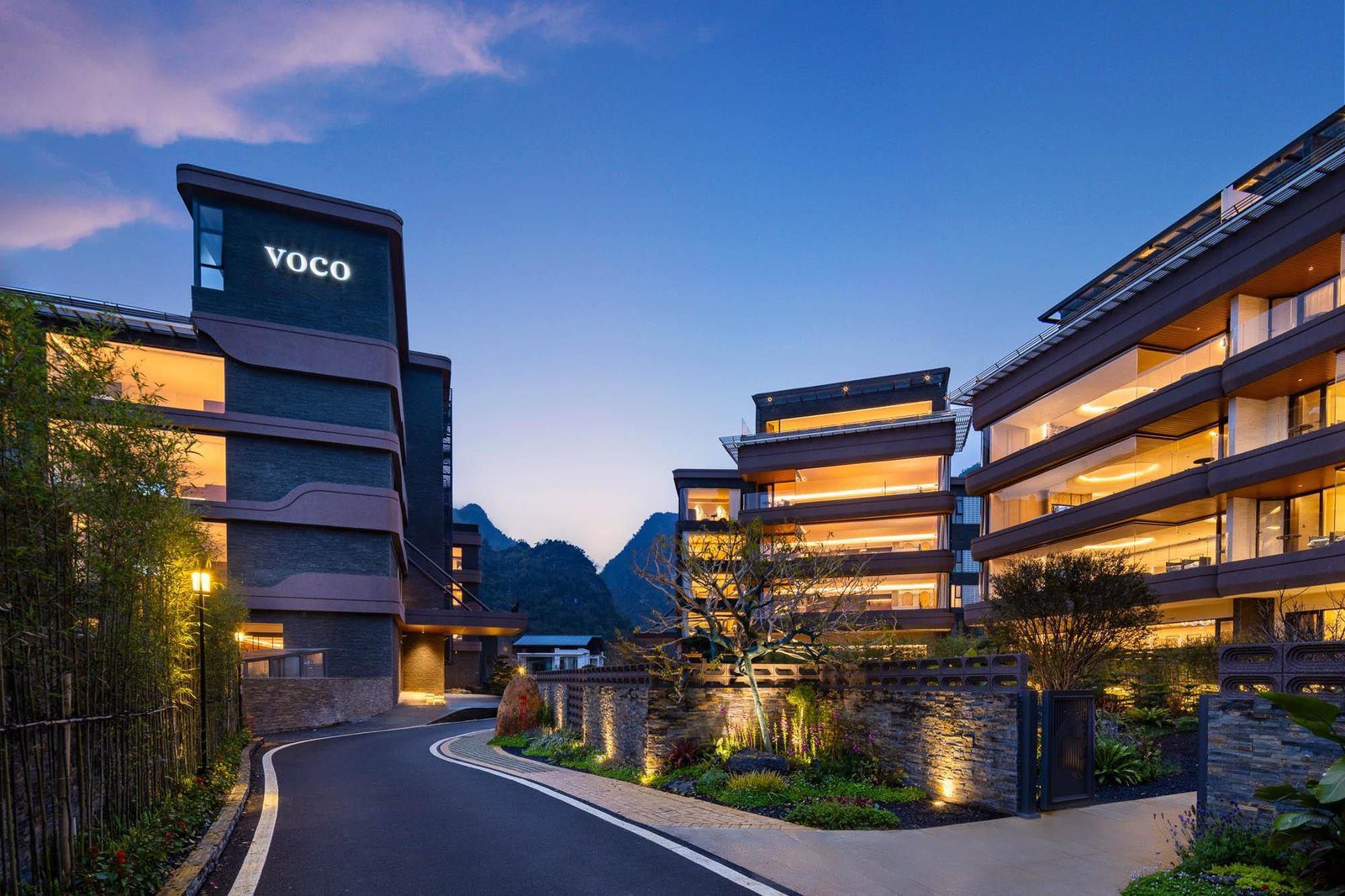 傍晚的桂林陽朔 voco 酒店入口車道，左側塔樓亮著 VOCO 字樣，右側是石牆花圃與整排落地窗的客房樓