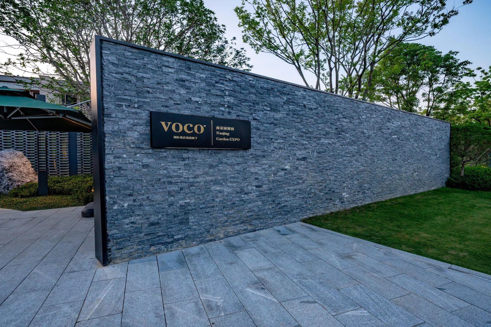南京園博園voco酒店入口的深灰色石牆，掛著 VOCO 與 Nanjing Garden EXPO 字樣的招牌，後方是綠樹