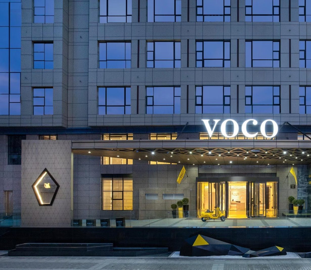 傍晚西安經開 voco 酒店的入口，雨遮上方亮著 VOCO 招牌，旋轉門內透出黃色燈光，門口停著一台黃色機車，前方有黑黃相間的幾何石雕