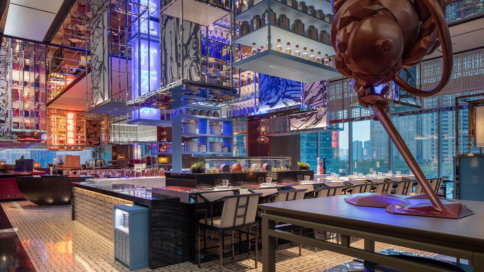 成都 W 酒店型樂特色餐廳的開放式主廚工作檯：中央金屬料理檯配高腳椅，上方懸掛酒瓶與器皿層架，背景是藍色霓虹與落地窗外的高樓