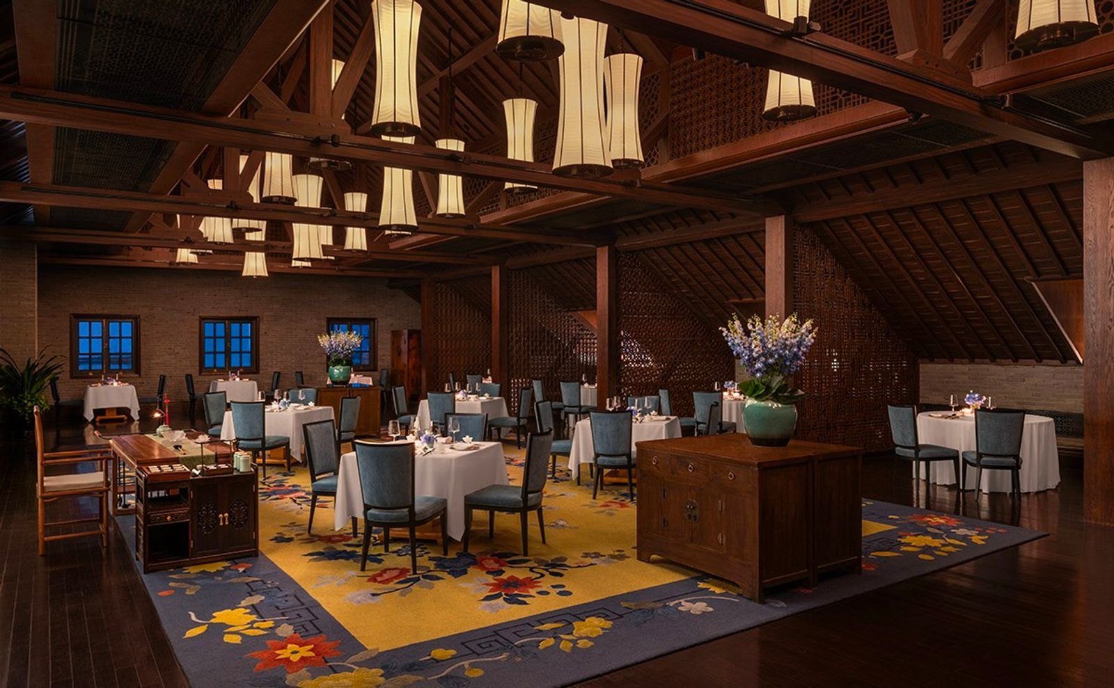 上海外灘華爾道夫酒店蔚景閣中餐廳，斜頂木梁下掛著成排白色燈籠，鋪白桌巾的餐桌擺在黃色花卉地毯上，牆邊是小窗