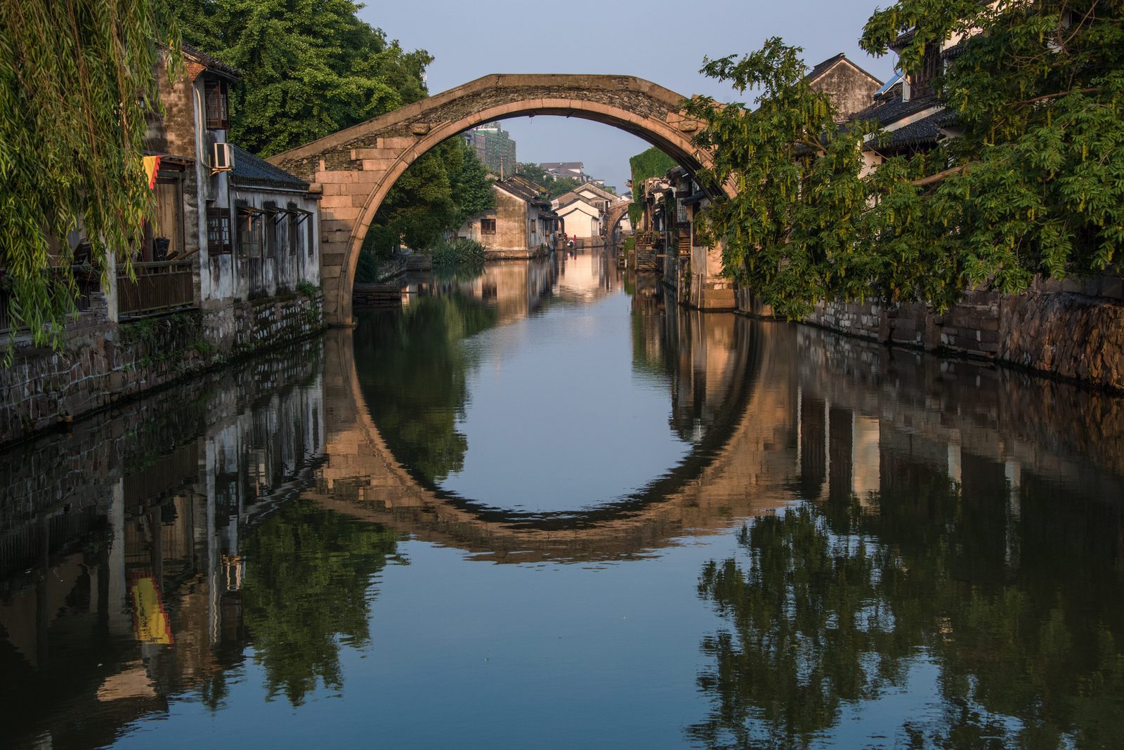 江南水鄉古鎮河道上的單孔石拱橋,橋洞與水中倒影連成圓形,兩岸有老屋與綠樹