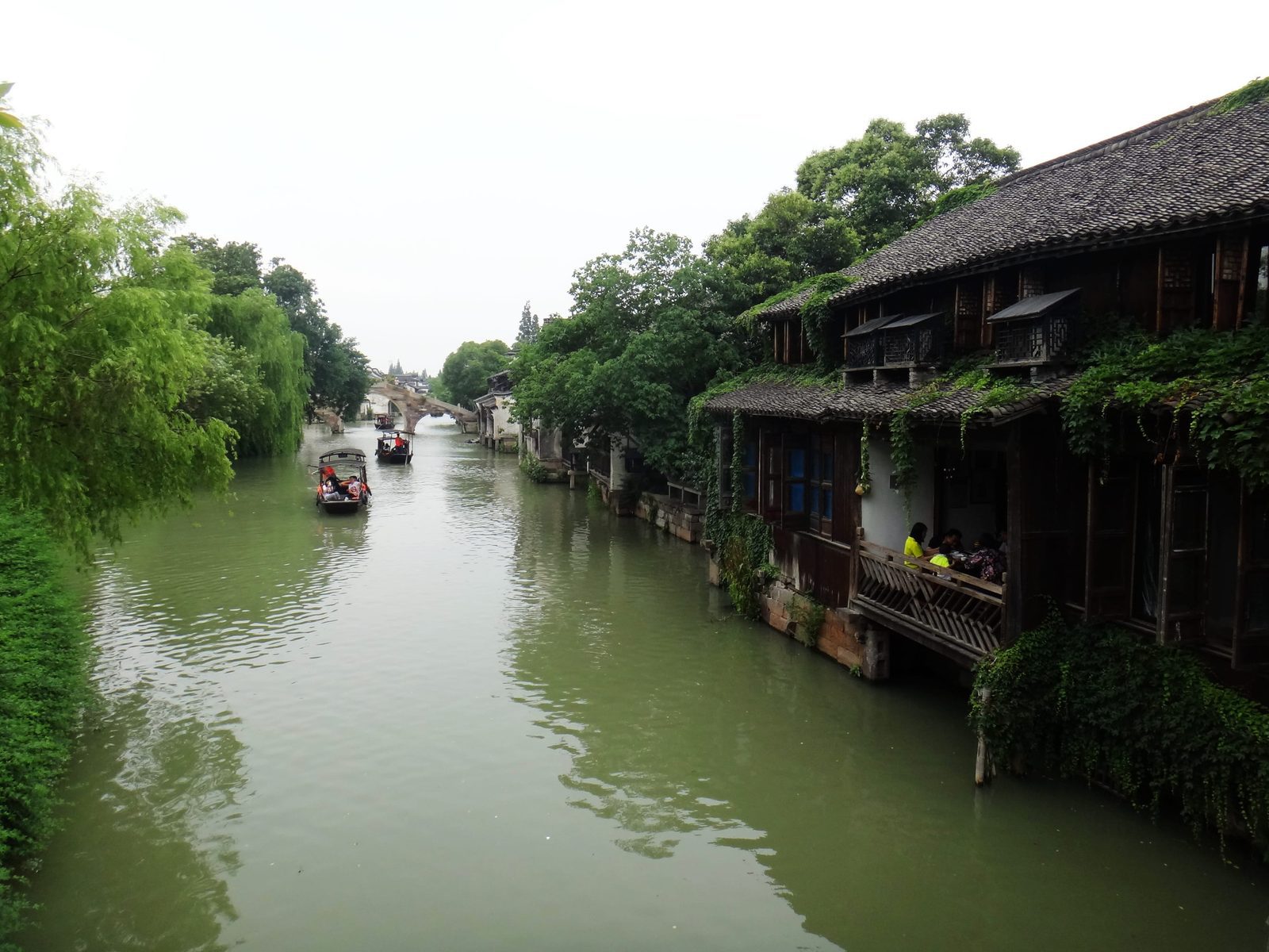 江南水鄉古鎮寬闊的河道,遠處有石拱橋,右岸是攀滿藤蔓的木樓,河上有小船