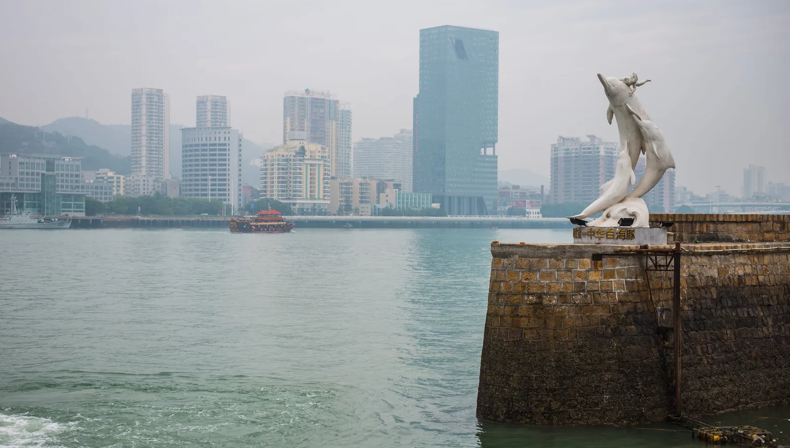 陰天霧氣中的海港,岸邊石堤上有海豚雕像,對岸是城市高樓