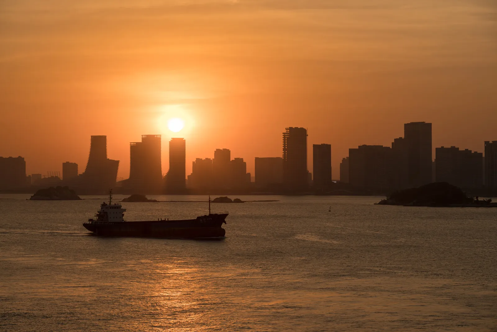 夕陽落在海港城市天際線後方,海面上有貨船駛過